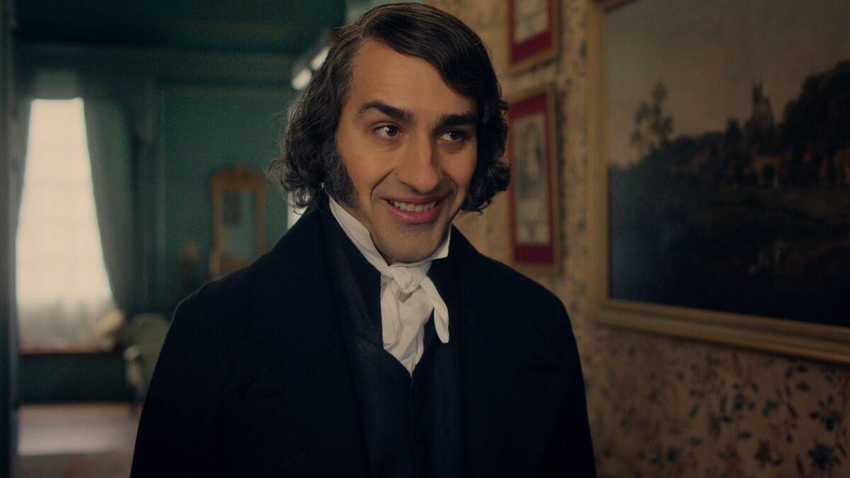 "A man in early 19th-century attire with sideburns smiles in a warmly lit, vintage room with floral wallpaper, framed paintings, and a window in the background."