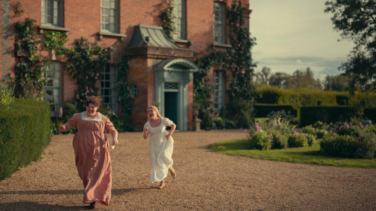 "Two women in period dresses run joyfully on a gravel path in front of a large brick house with ivy and flowers, surrounded by hedges and gardens under a partly cloudy sky."
