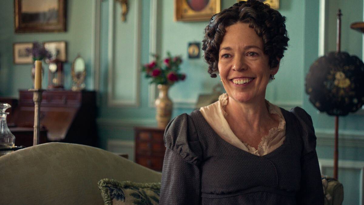 "A smiling woman in period costume sits on a couch in a vintage, elegant living room with teal walls, antique furniture, framed art, and a flower vase in the background."