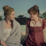 "Two women in historical clothing sit outside on a rooftop, smiling at each other with greenery and blue sky in the background. Pillows and a cup are nearby, creating a relaxed, intimate setting."