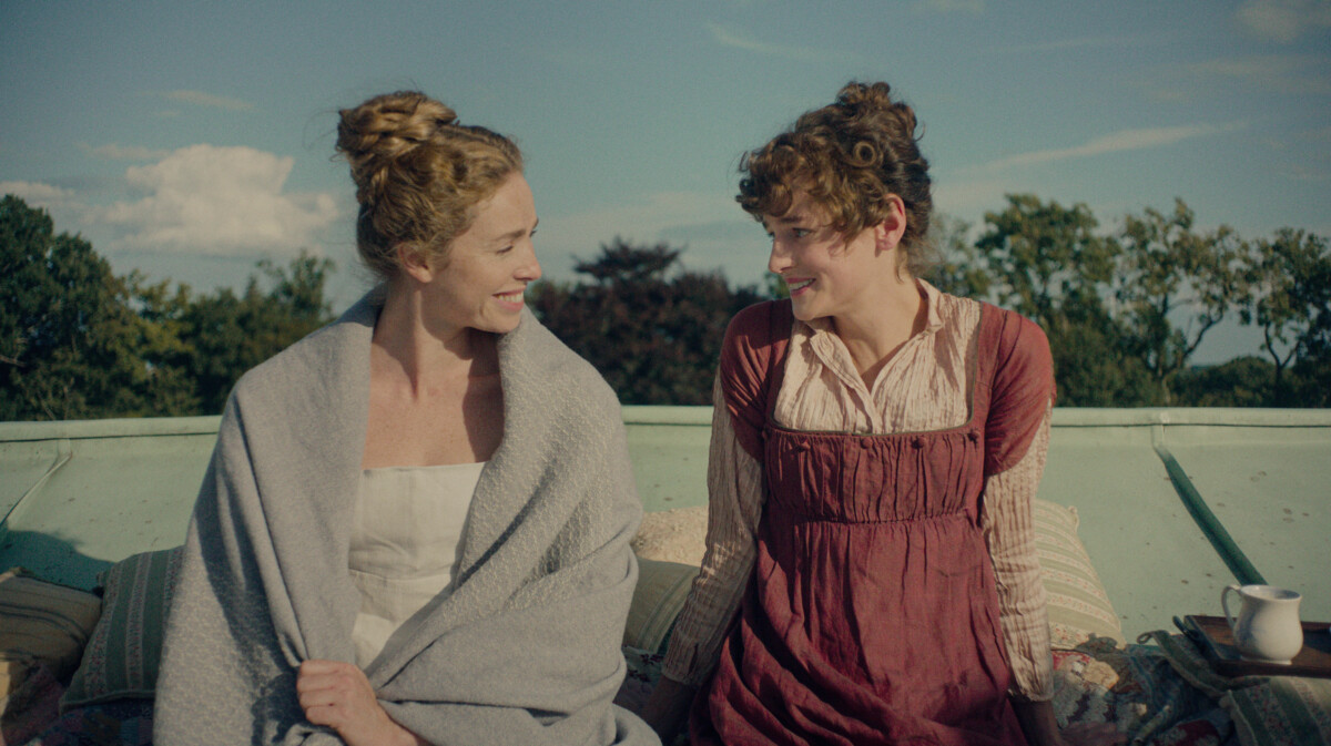 "Two women in historical clothing sit outside on a rooftop, smiling at each other with greenery and blue sky in the background. Pillows and a cup are nearby, creating a relaxed, intimate setting."