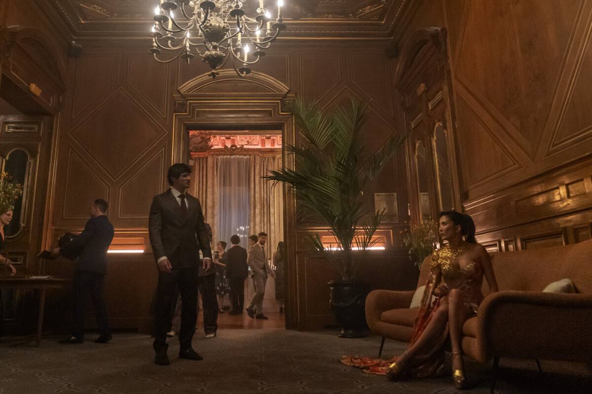 "A man in a suit stands in an elegant, wood-paneled lounge with a chandelier, while a woman in a gold dress sits on a couch; other people are visible in the background socializing at a formal event."