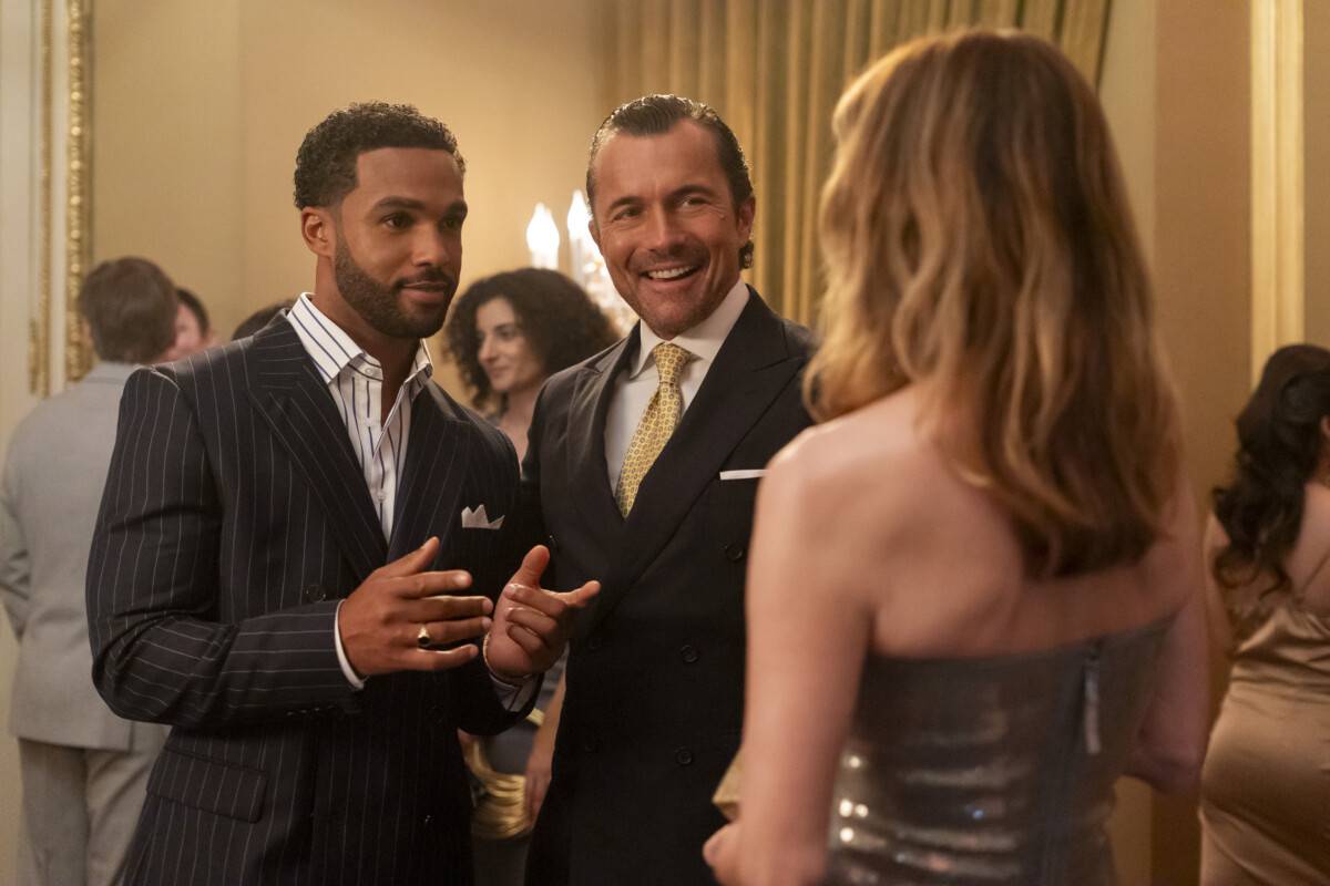 "Two men in suits and a woman in a metallic dress converse at an elegant indoor party, surrounded by other formally dressed guests in a well-lit, upscale setting with gold accents and curtains."