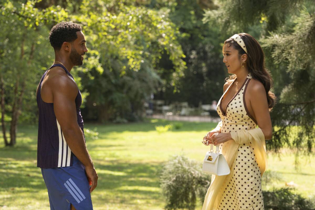 "A man in athletic wear and a woman in a yellow polka dot dress stand facing each other in a sunlit park surrounded by trees, engaged in conversation on a summer day."